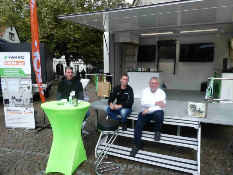 Drei Männer sitzen vor dem mobilen Ausstellungsfahrzeug von FAKRO und KRONmat während einer Produktpräsentation im Freien.