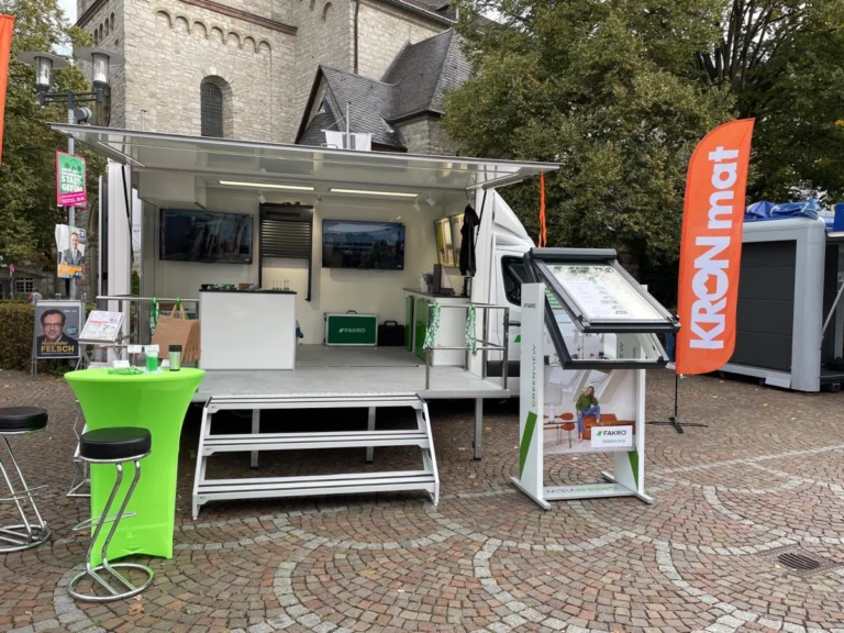 Präsentationsfahrzeug von FAKRO und KRONmat mit Infomaterialien, Dachfenster-Modell und Messestand auf einem Platz vor einer Kirche.